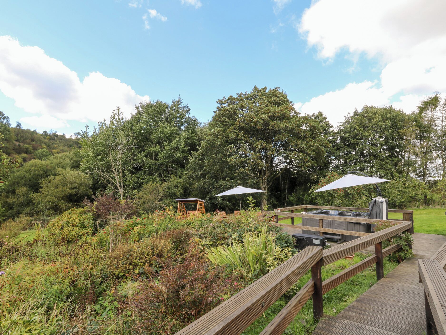 Une terrasse en bois avec un jacuzzi, des tables de pique-nique et des parasols, le tout dans un cadre extérieur luxuriant et verdoyant.