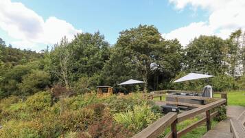 Une terrasse en bois avec un jacuzzi, des tables de pique-nique et des parasols, le tout dans un cadre extérieur luxuriant et verdoyant.