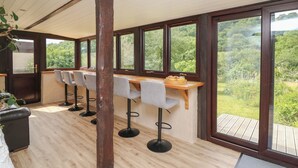 A dining area with a long wooden table and bar stools, large windows with a view of greenery, and a wooden ceiling. - Penarth (Llandrindod Wells)