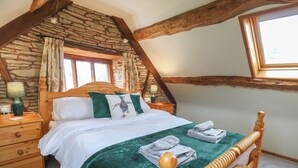 A bedroom with a stone wall, wooden beams, a wooden bed frame, a green bedspread, and a window with curtains. - Penarth (Llandrindod Wells)