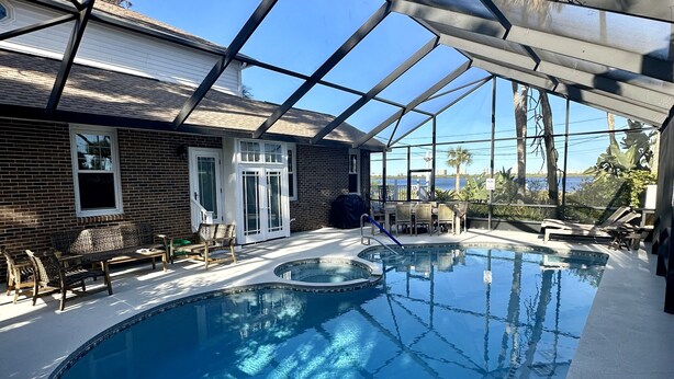 A covered swimming pool with a clear view of the surrounding area, including a building with a brick facade and a door, and a view of palm trees and a body of water in the background. - Otium Villa - Waterfront, intercoastal villa with a Spa-like feel. (Port Orange)