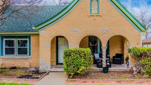 A two-story brick house with a green roof, a front porch with outdoor furniture, and a well-maintained garden. - Cozy Craftsman in the Center of Oak Cliff (Dallas)