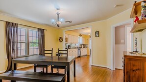 A dining area with a wooden table and chairs, a kitchen in the background, and a hallway leading to another room. - Cozy Craftsman in the Center of Oak Cliff (Dallas)