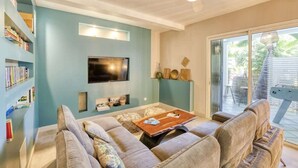 A living room with a sofa set, a coffee table, a flat-screen TV mounted on the wall, and a bookshelf filled with books.