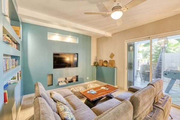 A living room with a sofa set, a coffee table, a flat-screen TV mounted on the wall, and a bookshelf filled with books.