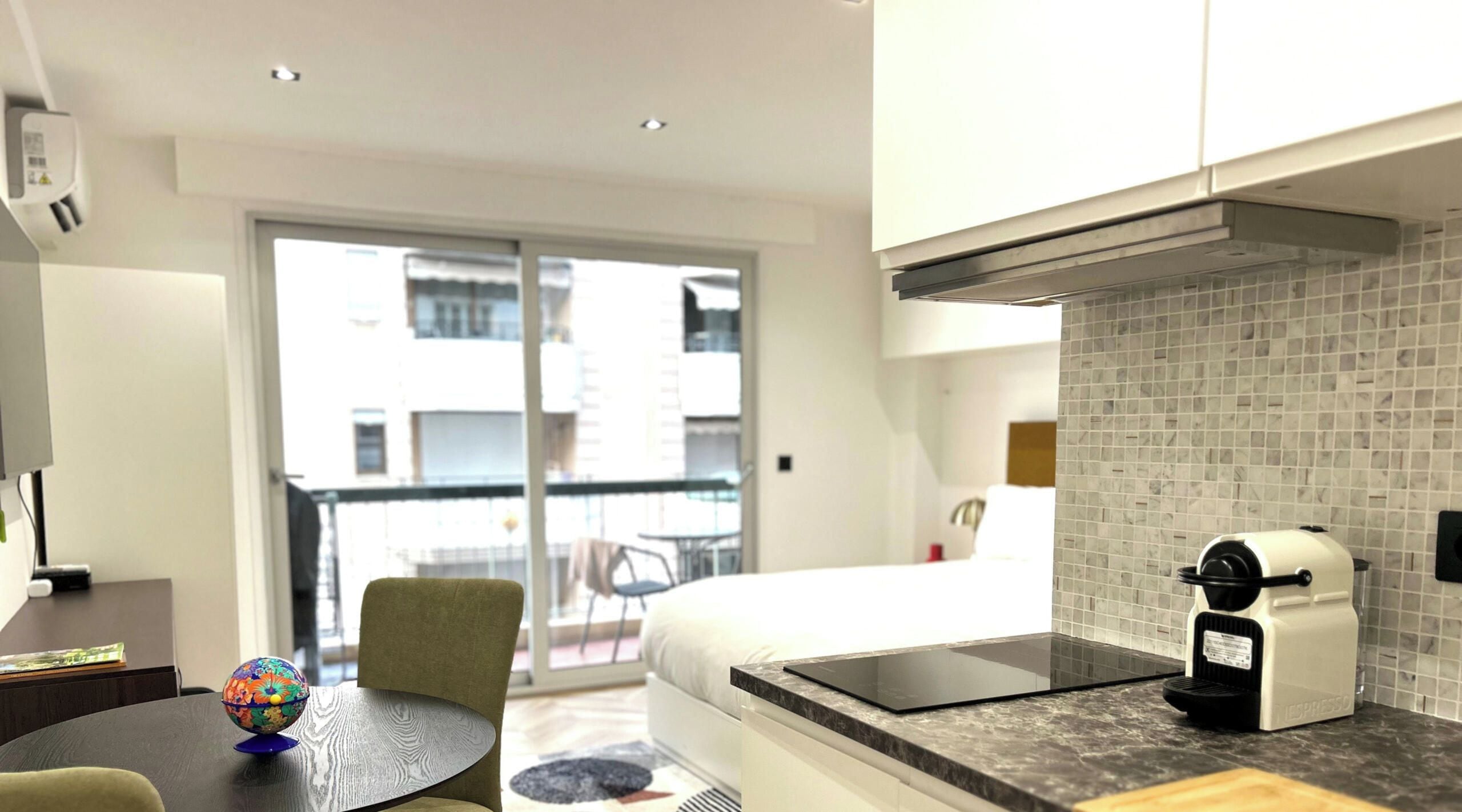 Ein modernes Hotelzimmer mit einem Bett, einem kleinen Tisch mit einem Globus, einer Kaffeemaschine und einem Balkon mit Blick auf Gebäude.