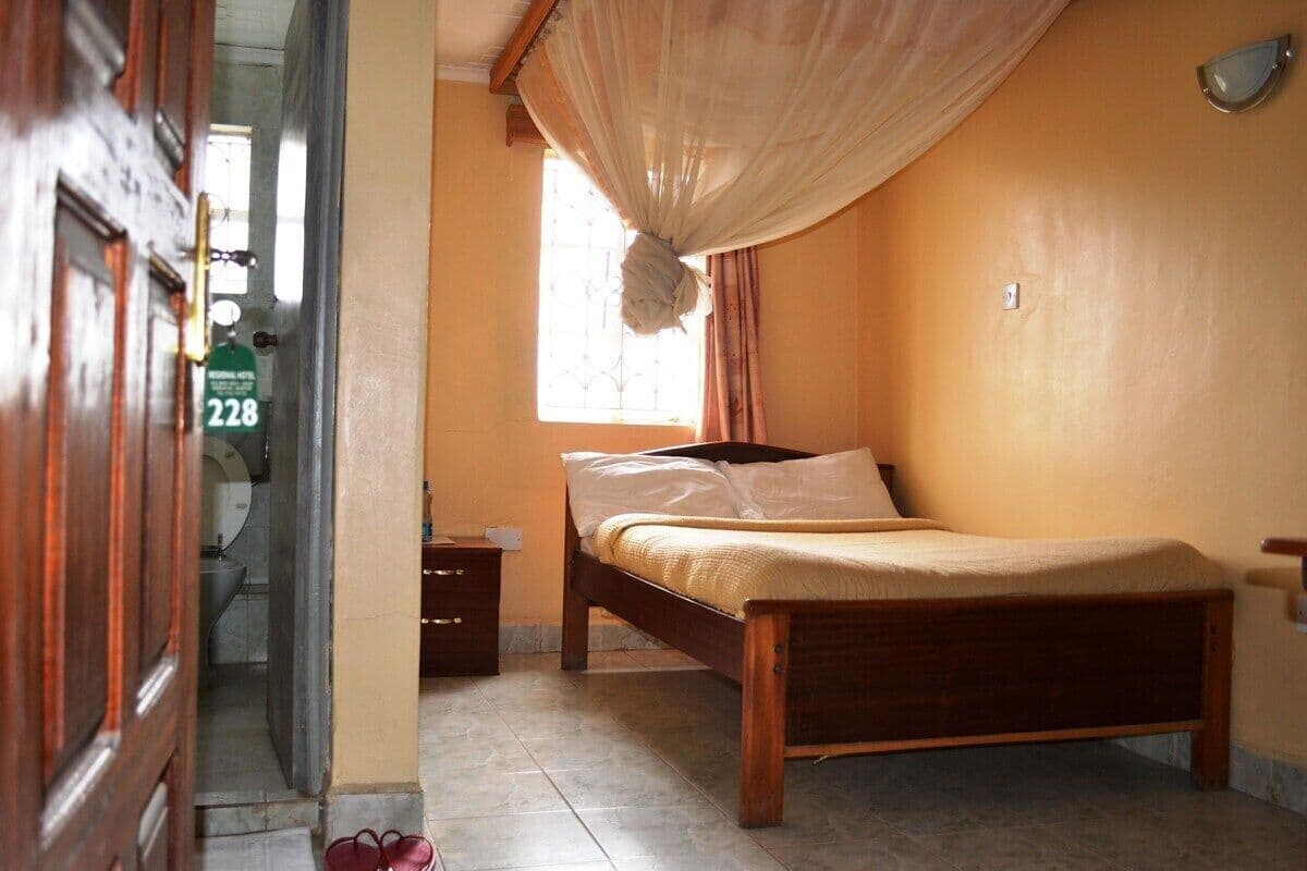 A bedroom with a wooden bed, a window with curtains, a nightstand, and a bathroom visible through an open door.