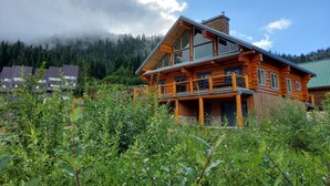 A log cabin with a balcony, surrounded by lush greenery and mountains in the background. - Bear Behind Lodge (Fraser Valley C)