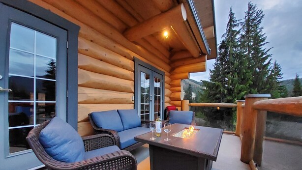 A log cabin with a wooden exterior, a glass door, a seating area with wicker furniture, and a fire feature on the patio. - Bear Behind Lodge (Fraser Valley C)