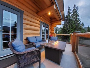 A log cabin with a wooden exterior, a glass door, a seating area with wicker furniture, and a fire feature on the patio. - Bear Behind Lodge (Fraser Valley C)