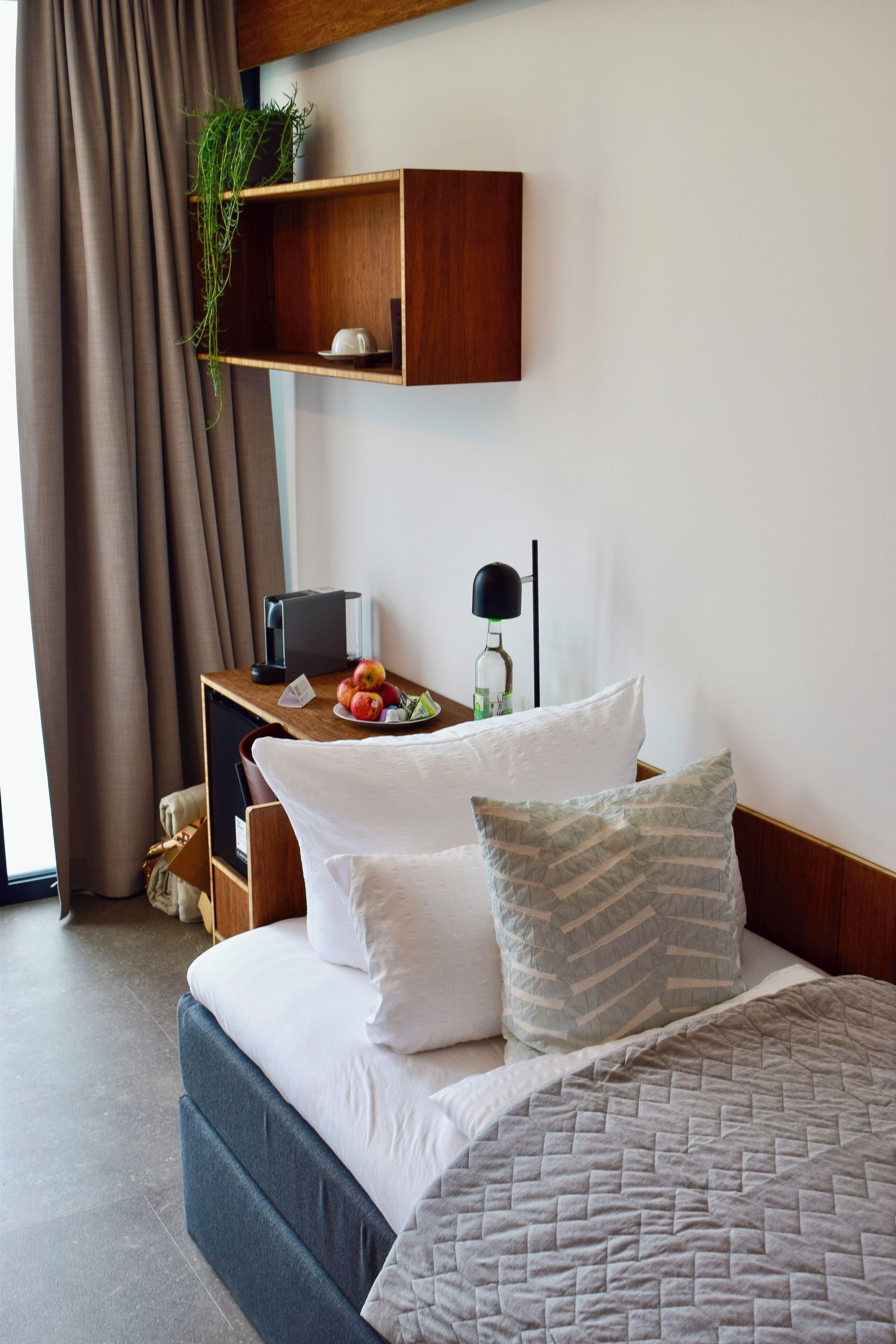Una habitación de hotel con una cama, un escritorio con ordenador, una lámpara y un pequeño estante con una planta.