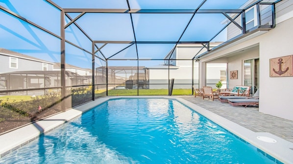 Una piscina con cerramiento de vidrio, mobiliario exterior y vistas a casas vecinas.