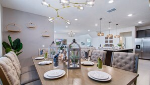 A modern dining area with a wooden table set for six, surrounded by tufted chairs. A chandelier hangs above, and the room features a kitchen in the background. - Windsor Island 5br Villa W/private Pool Spa Disney (Davenport)