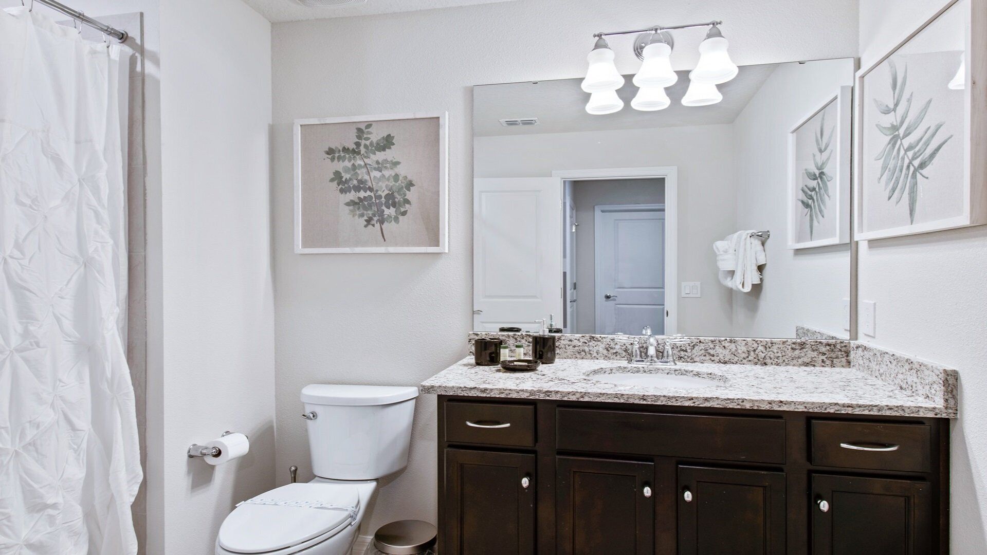 A bathroom with a white toilet, dark wooden cabinets, a granite countertop, and a large mirror with lights above.
