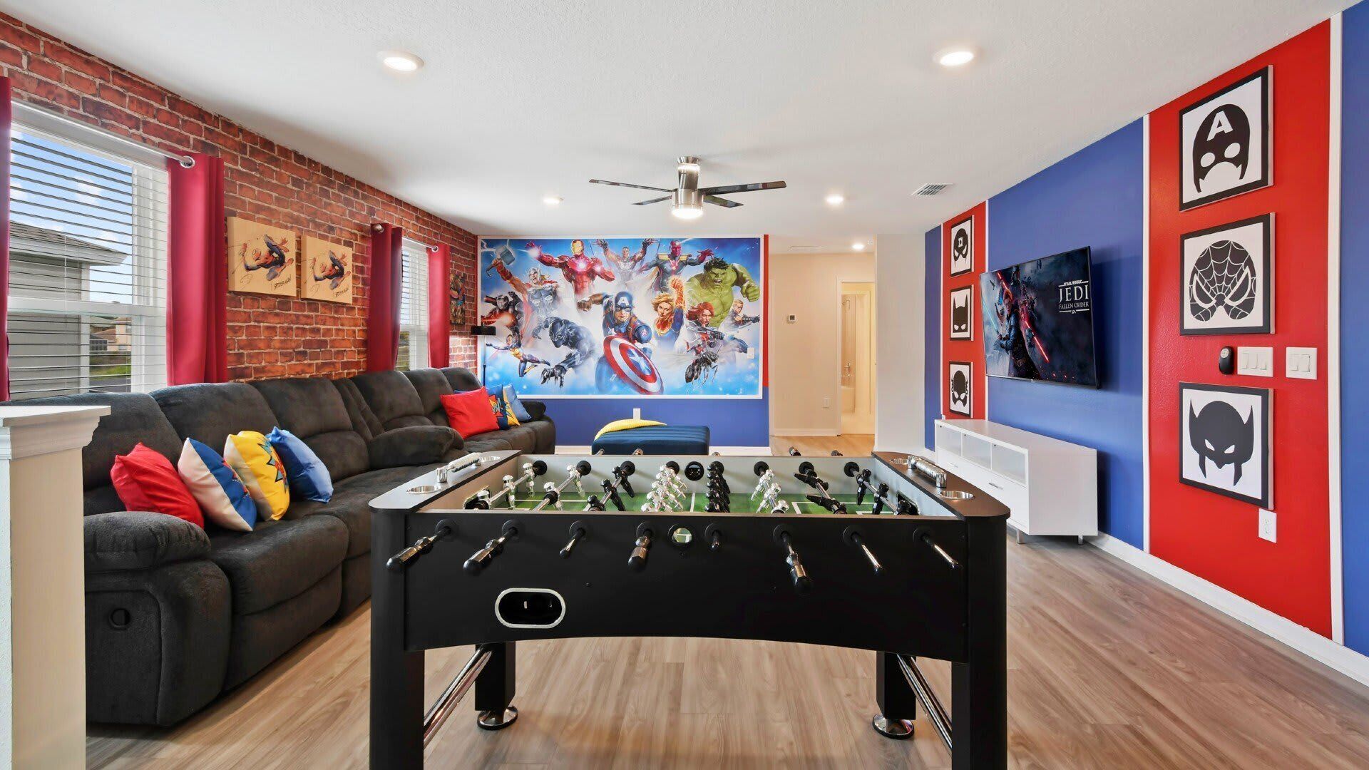 A living room with a foosball table, a sofa with colorful cushions, a large superhero-themed painting, and a TV mounted on a red wall.