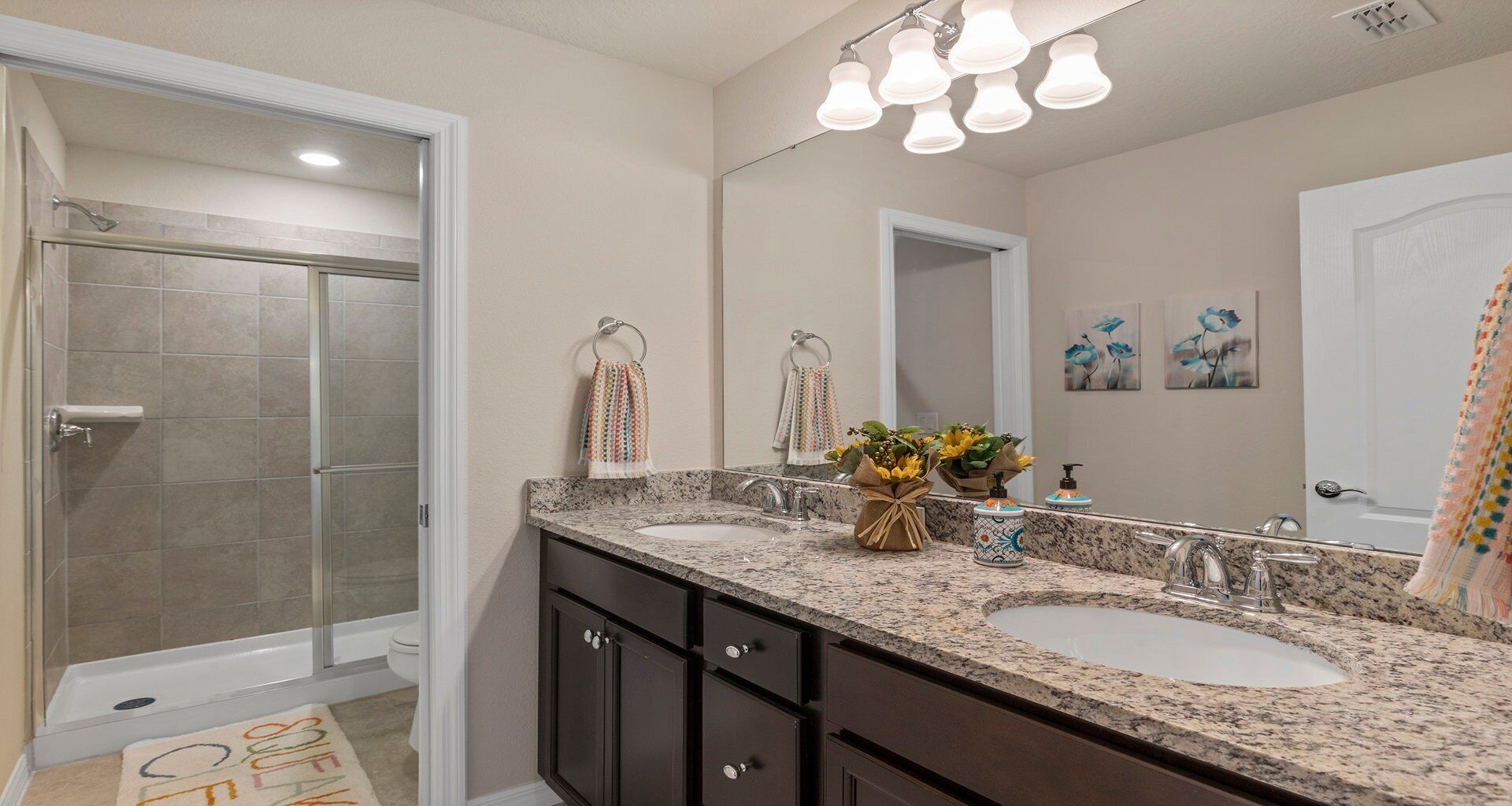 A bathroom with two sinks, a shower, and a large mirror. There are towels hanging on a rack and a small plant on the counter.