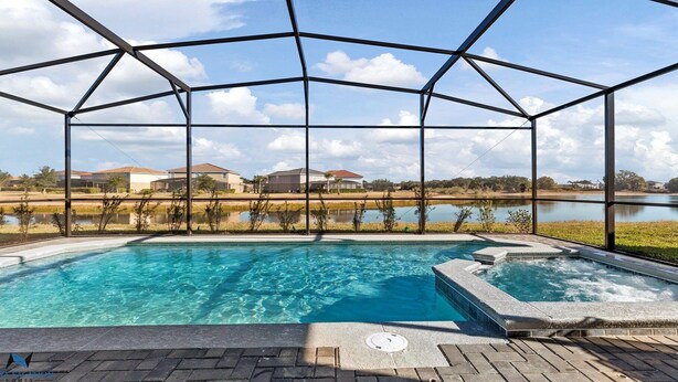 Ein Schwimmbad mit Glasüberdachung, umgeben von einer Ziegelterrasse und Blick auf Häuser und einen See.