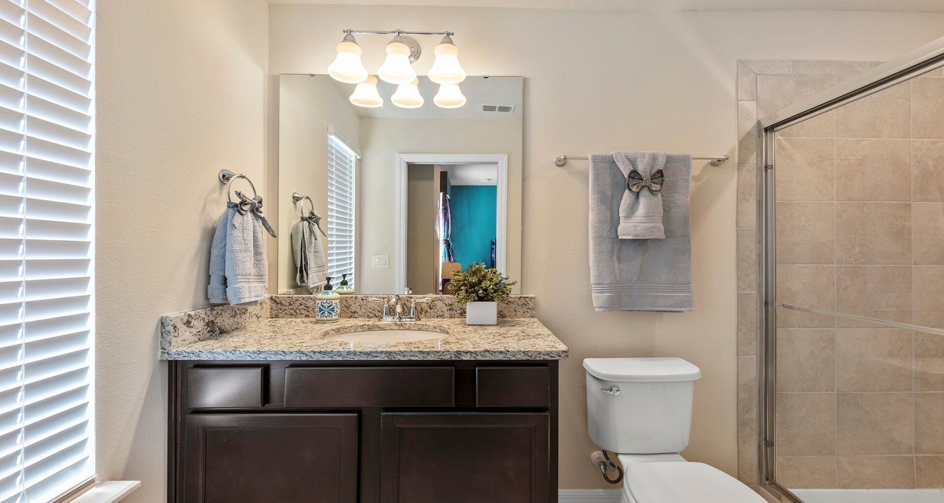 A bathroom with a granite countertop, a white toilet, a shower area, and a mirror with lights.
