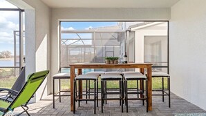 Eine Terrasse mit einem Holztisch und -stühlen, einem grünen Stuhl und einer Glastür, die einen Blick auf ein Haus und ein Gewässer freigibt.