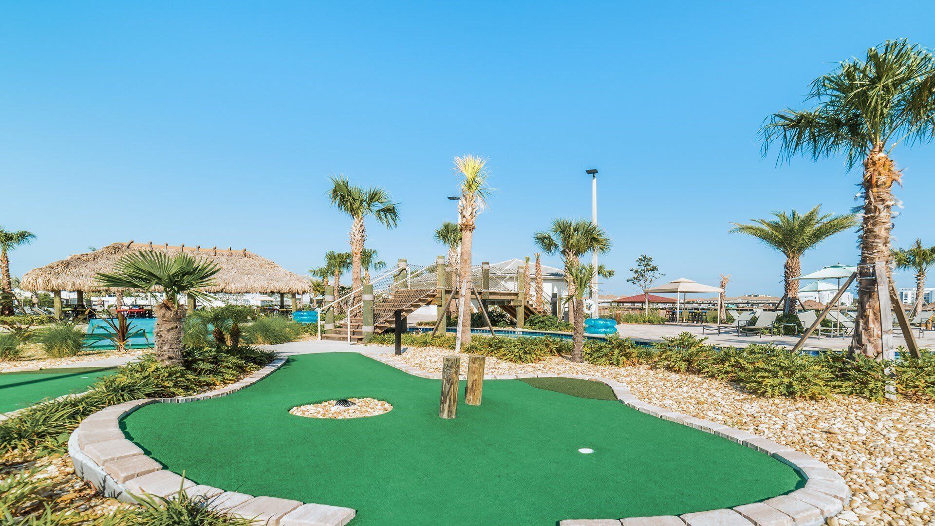 A miniature golf course with a green putting area, palm trees, and a thatched-roof structure.
