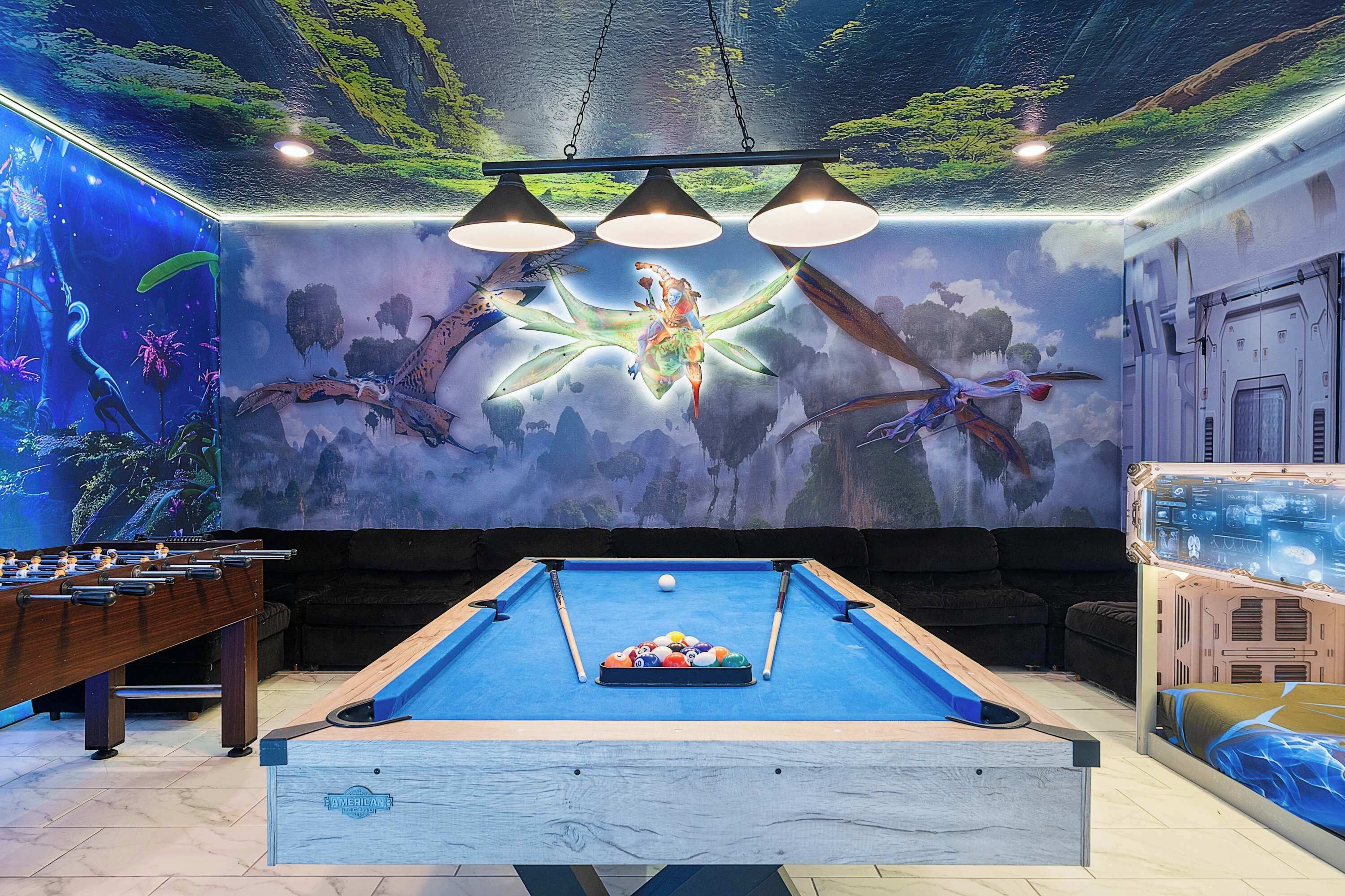 A pool table with a colorful ball rack, a foosball table, and a mural of a celestial scene with a figure on a starfish.