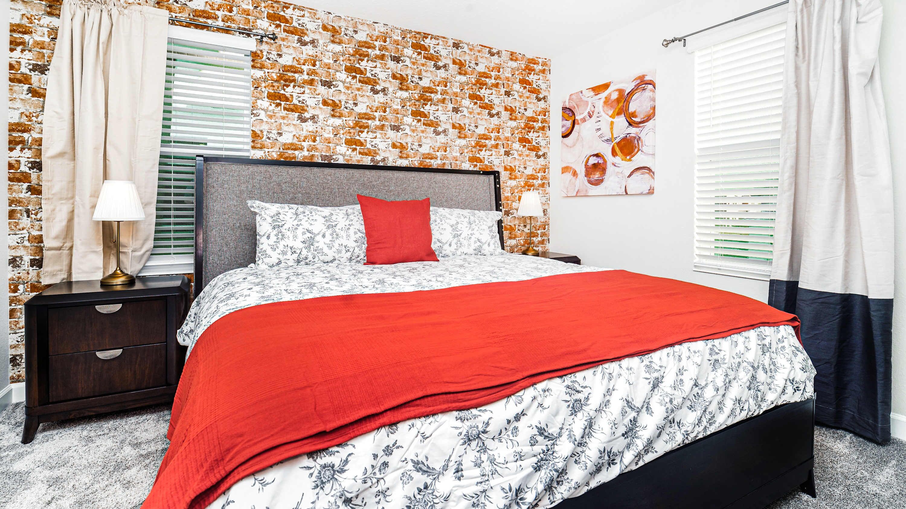 Een slaapkamer met een metselwerkmuur, een bed met een bloemenbedekking en rode kussens, een nachtkastje met een lamp, en een raam met jaloezieën.