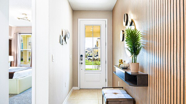 A hotel hallway with a white door, a small bench, a potted plant, and a wall-mounted shelf with decorative items.