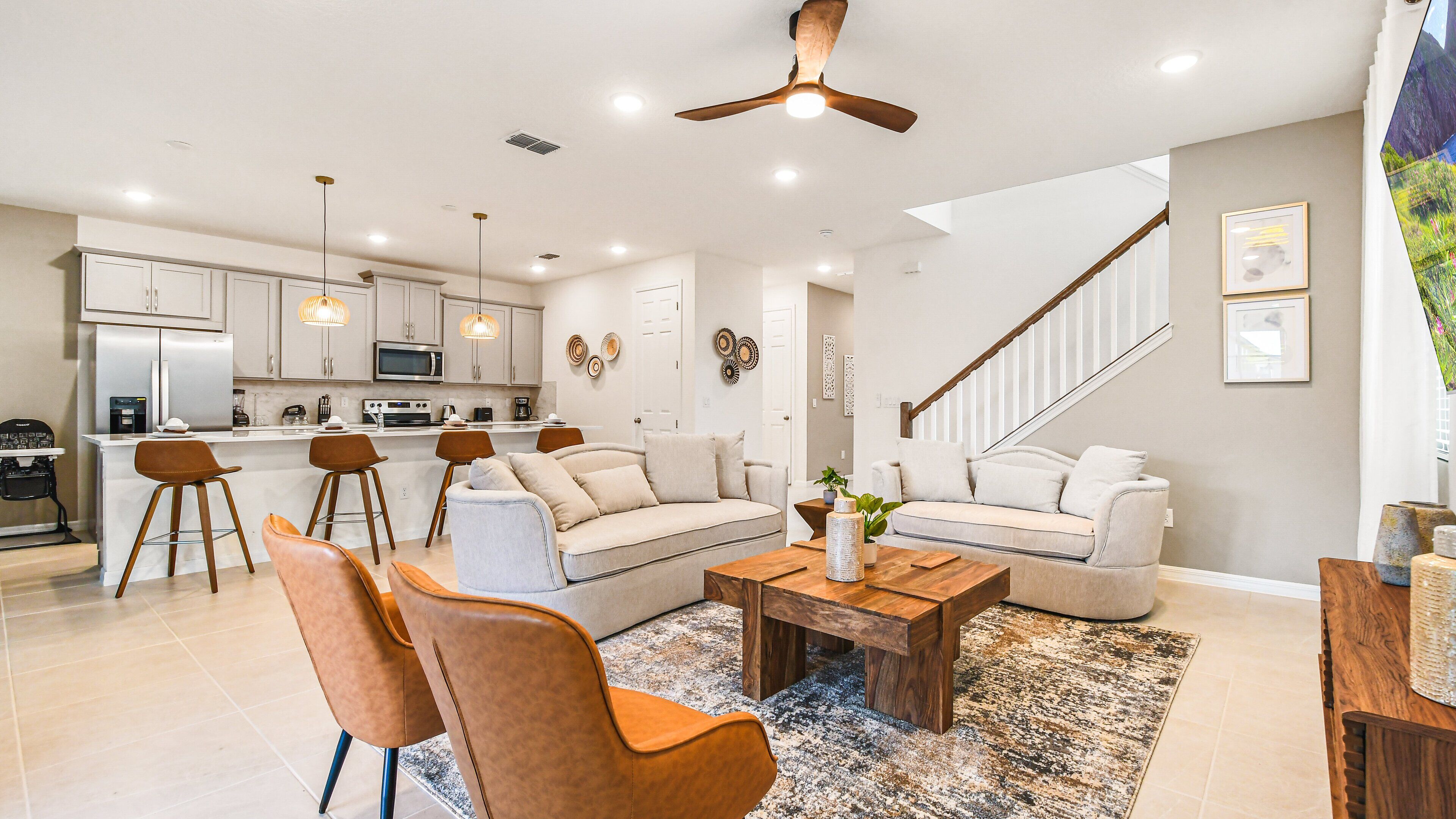 A modern kitchen with white cabinets, a central island, and bar stools. There is a living area with a sofa, coffee table, and chairs. A staircase with a wooden railing leads to another level.