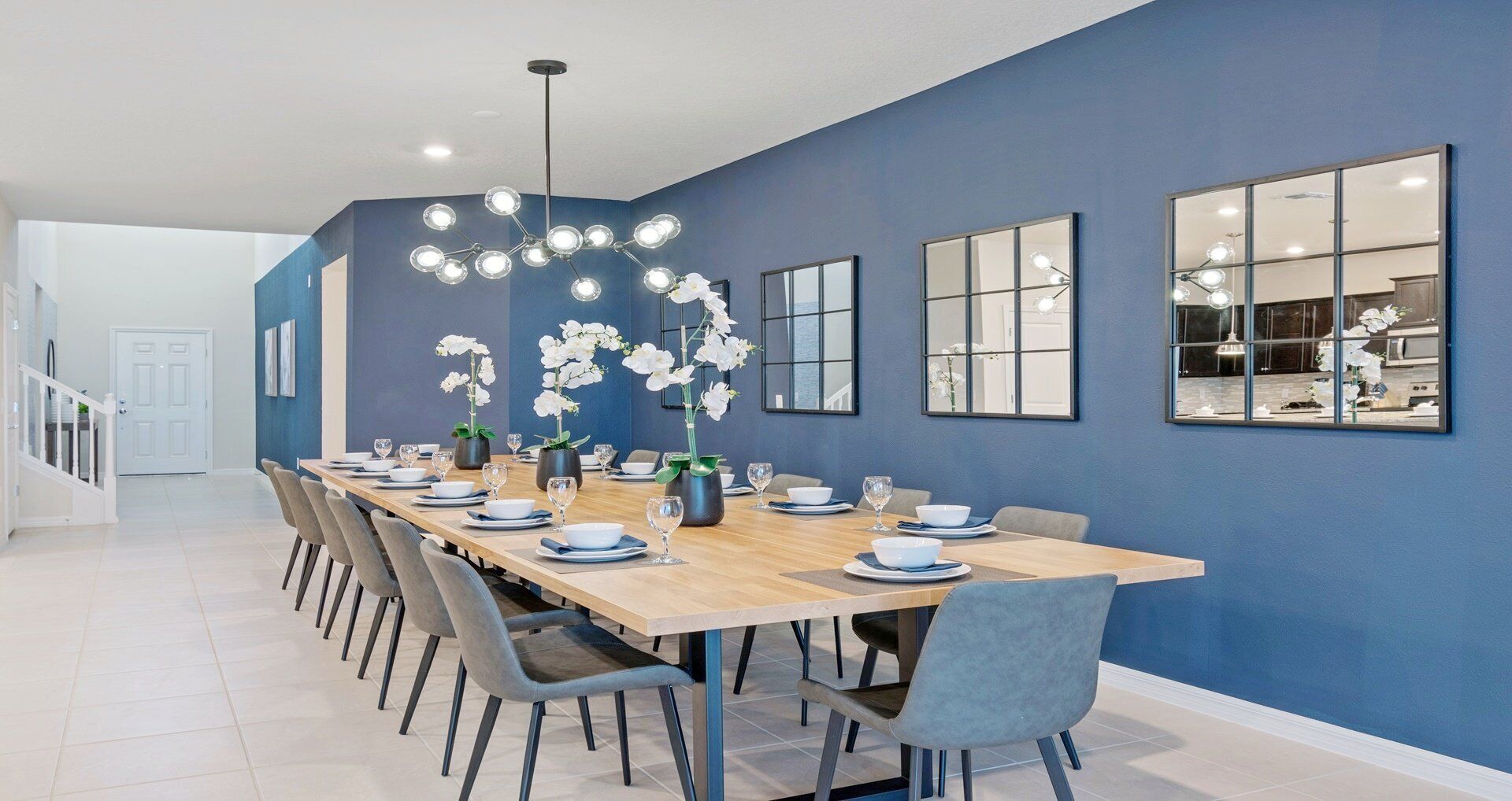 A modern dining area with a long table set for six, surrounded by grey chairs. There are white orchids in vases on the table, and a ceiling light fixture above.