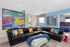A living room with a sectional sofa, colorful comic book-themed pillows, a blue blanket, and a red side table.