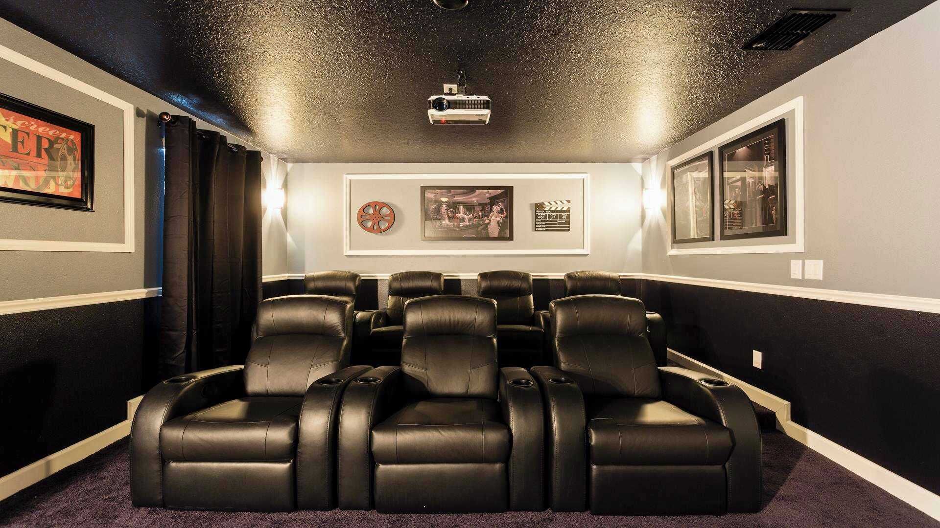 A home theater with six black leather reclining chairs, a ceiling-mounted projector, and framed movie posters on the walls.