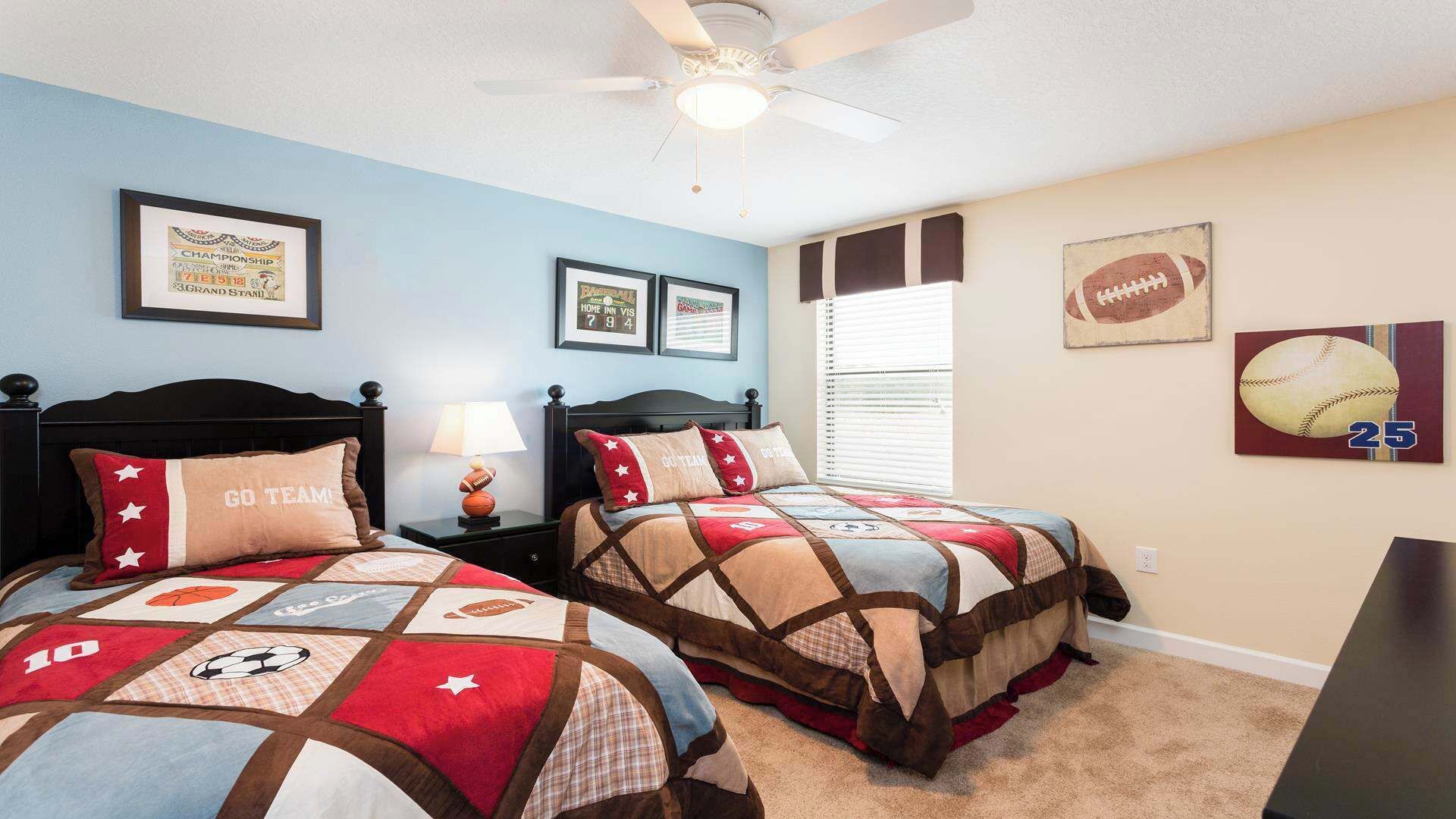 A bedroom with two beds, each with a sports-themed quilt, a ceiling fan, and framed sports memorabilia on the walls.