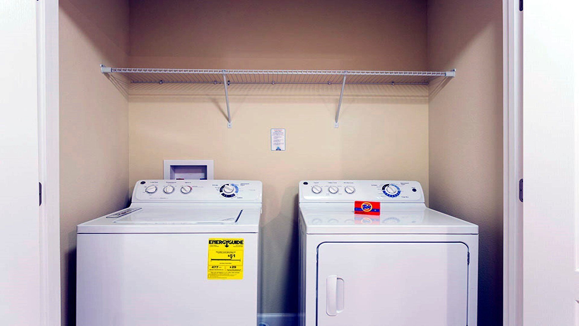 A laundry room with two stacked washers and dryers, a metal shelf above, and a power outlet on the wall.