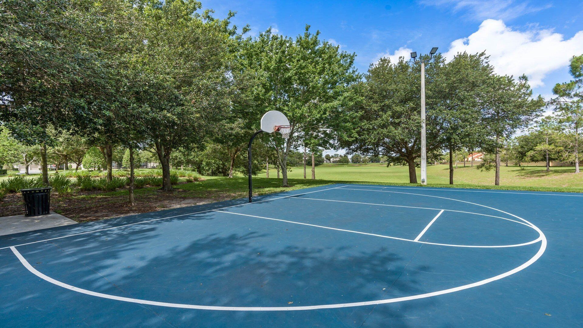 An outdoor basketball court with a hoop, surrounded by trees and a clear sky.