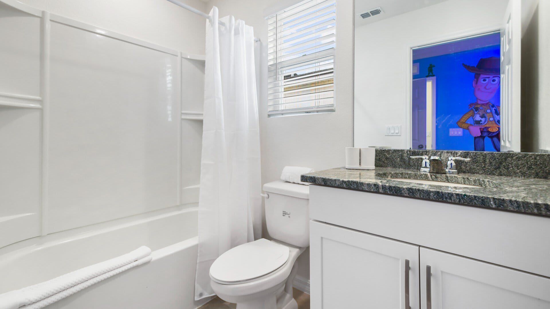 Un baño con inodoro blanco, ducha con cortina blanca, lavabo con encimera de granito y espejo que refleja un personaje de caricatura.