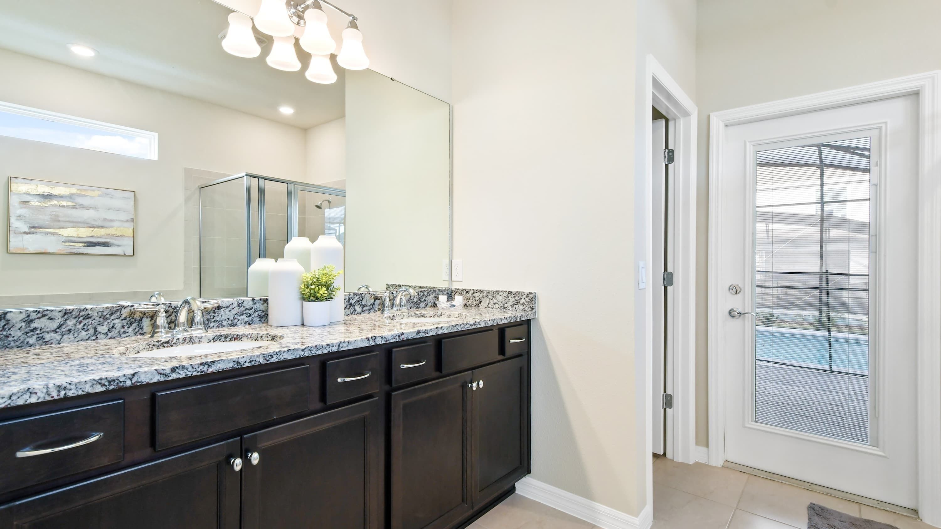 A modern bathroom with a double sink vanity, dark cabinetry, a large mirror, and a door leading to a balcony.