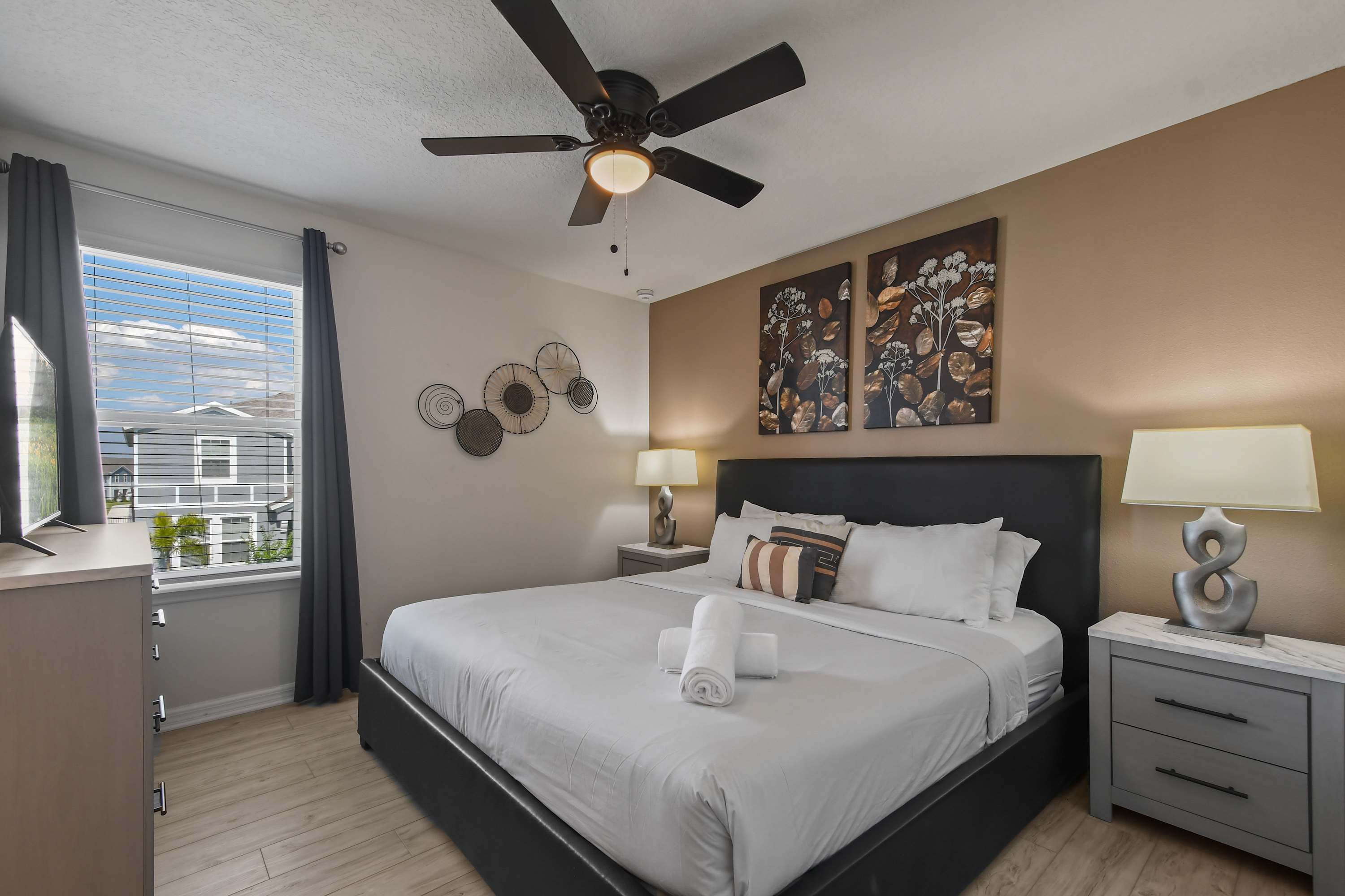 Ein modernes Hotelzimmer mit einem großen Bett, einem Deckenventilator, einem Fenster mit Vorhängen, einem Nachttisch mit Lampe und Kunstwerken an den Wänden.