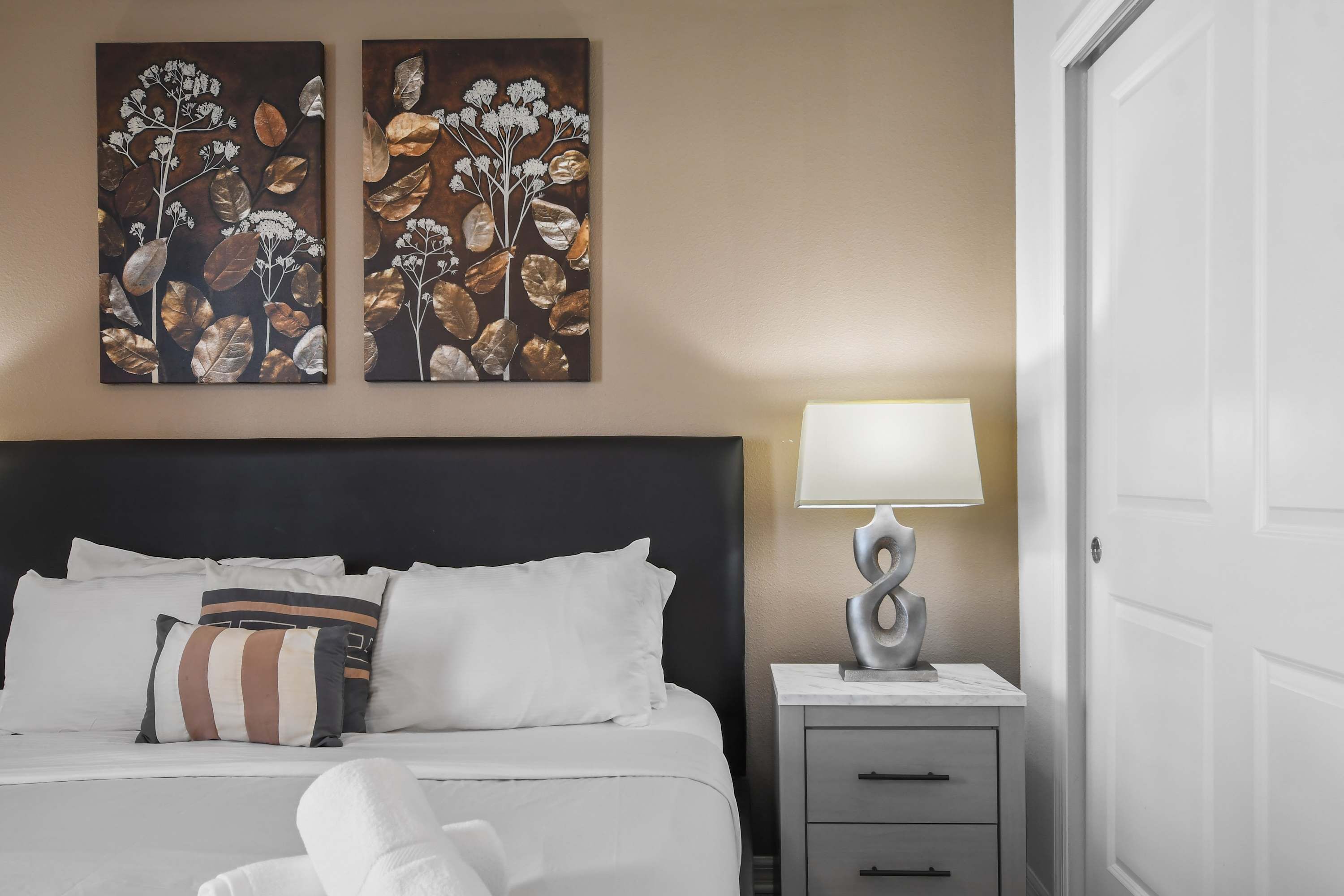 Un lit bien fait, avec une literie blanche et des oreillers à rayures, une table de chevet avec une lampe, et deux œuvres d’art accrochées au mur représentant des motifs floraux.