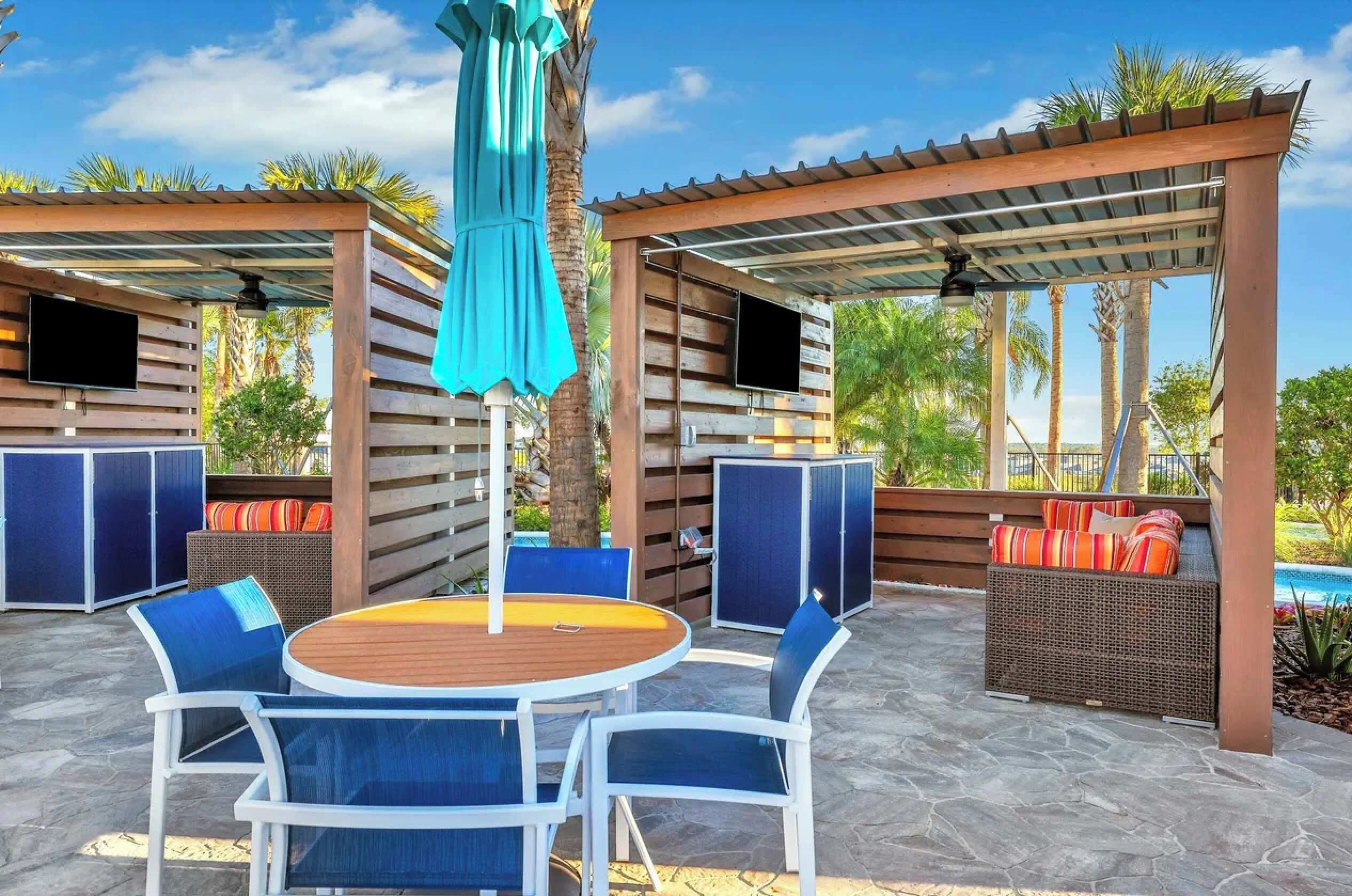 Un patio al aire libre con una mesa redonda de madera y sillas azules, una zona de estar cubierta con mobiliario de mimbre y un televisor montado en una pared de madera.