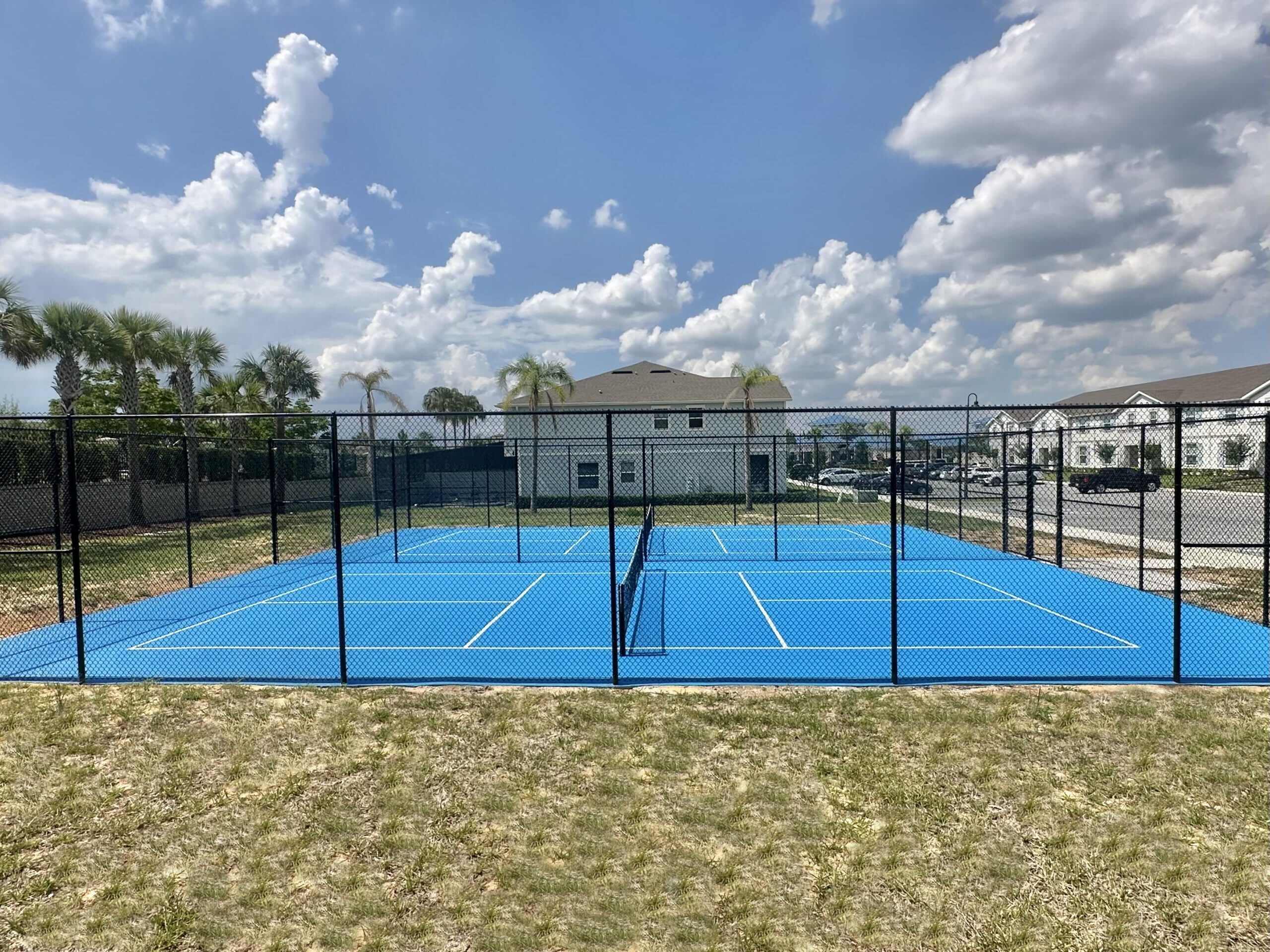 Una cancha de tenis al aire libre rodeada por una cerca negra, con un edificio y palmeras al fondo.