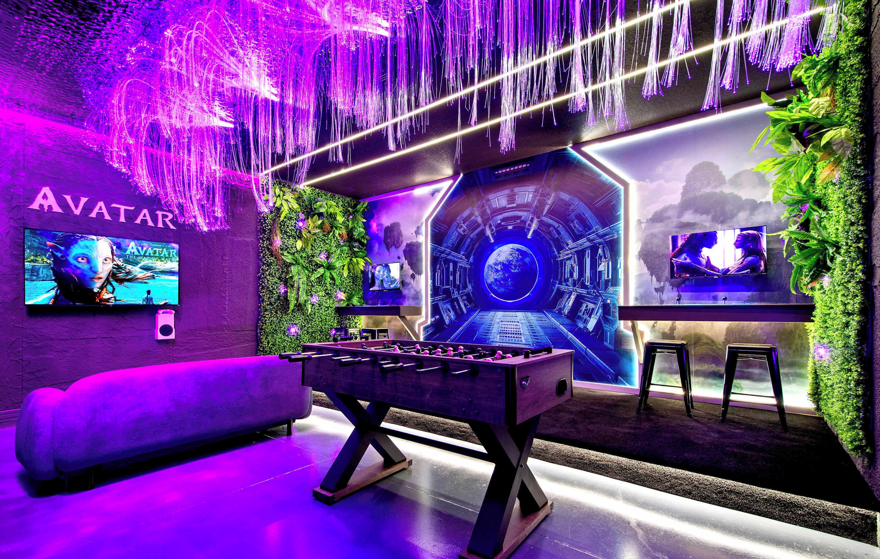A room with a foosball table, a sofa, and two TVs on the wall.