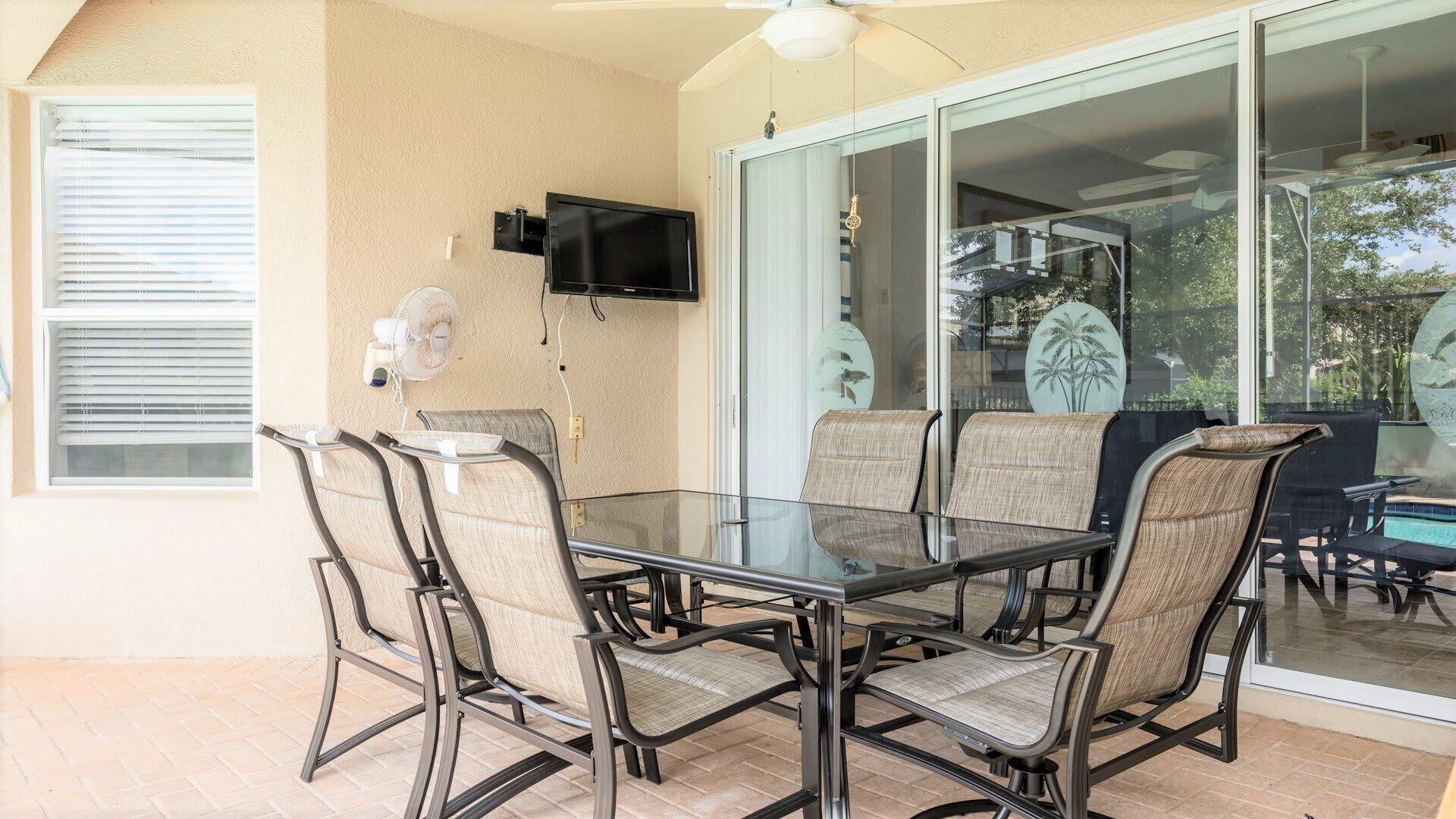Une table à plateau en verre, quatre chaises, un ventilateur de plafond et un téléviseur fixé au mur, le tout dans une véranda dotée d’une porte-fenêtre coulissante.