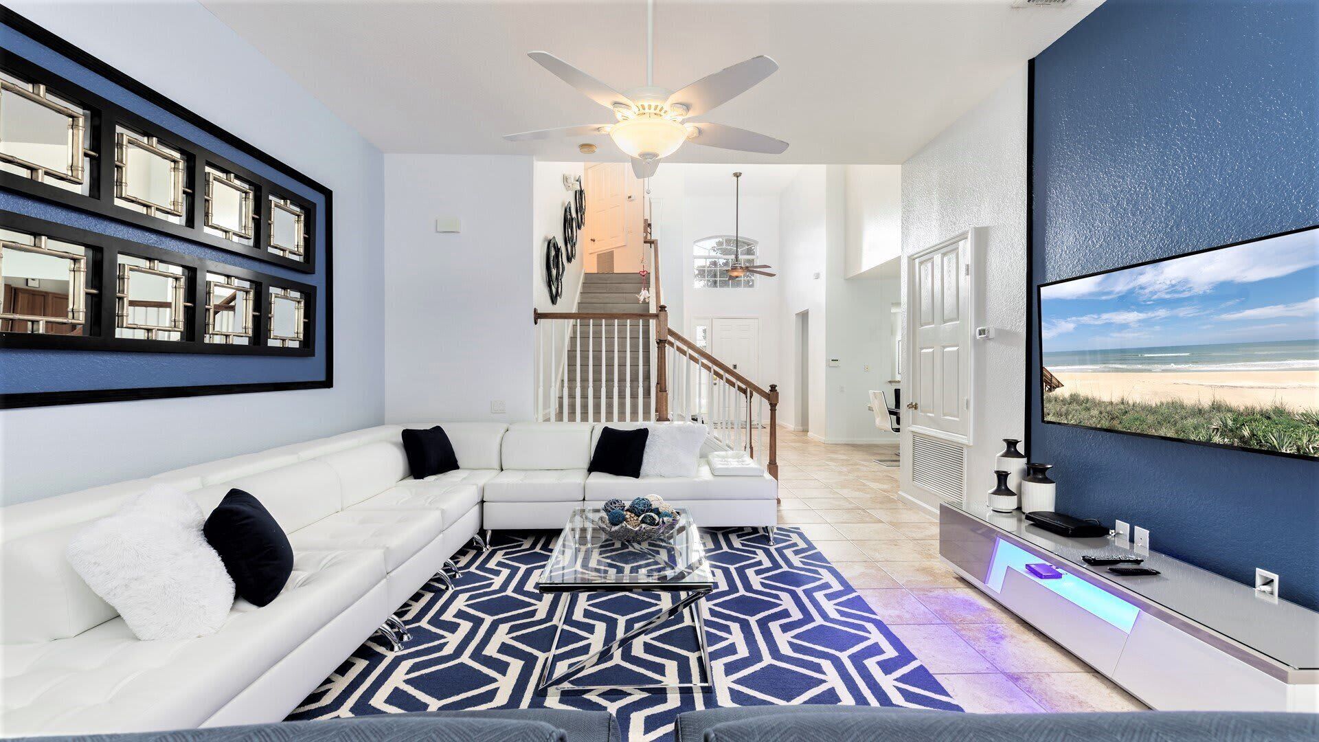 A modern living room with a white sofa, a coffee table, and a patterned rug. There is a staircase leading to another level, and a large TV screen displaying a beach view.