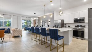 A modern kitchen with white cabinets, stainless steel appliances, and blue bar stools. - Bella Vida 12br Kids World With Pool Spa Near Disney 4497 (Kissimmee)