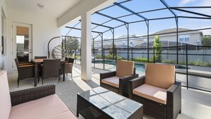 A covered outdoor area with wicker furniture, a glass table, and a view of a swimming pool and houses in the background. - Bella Vida 12br Kids World With Pool Spa Near Disney 4497 (Kissimmee)