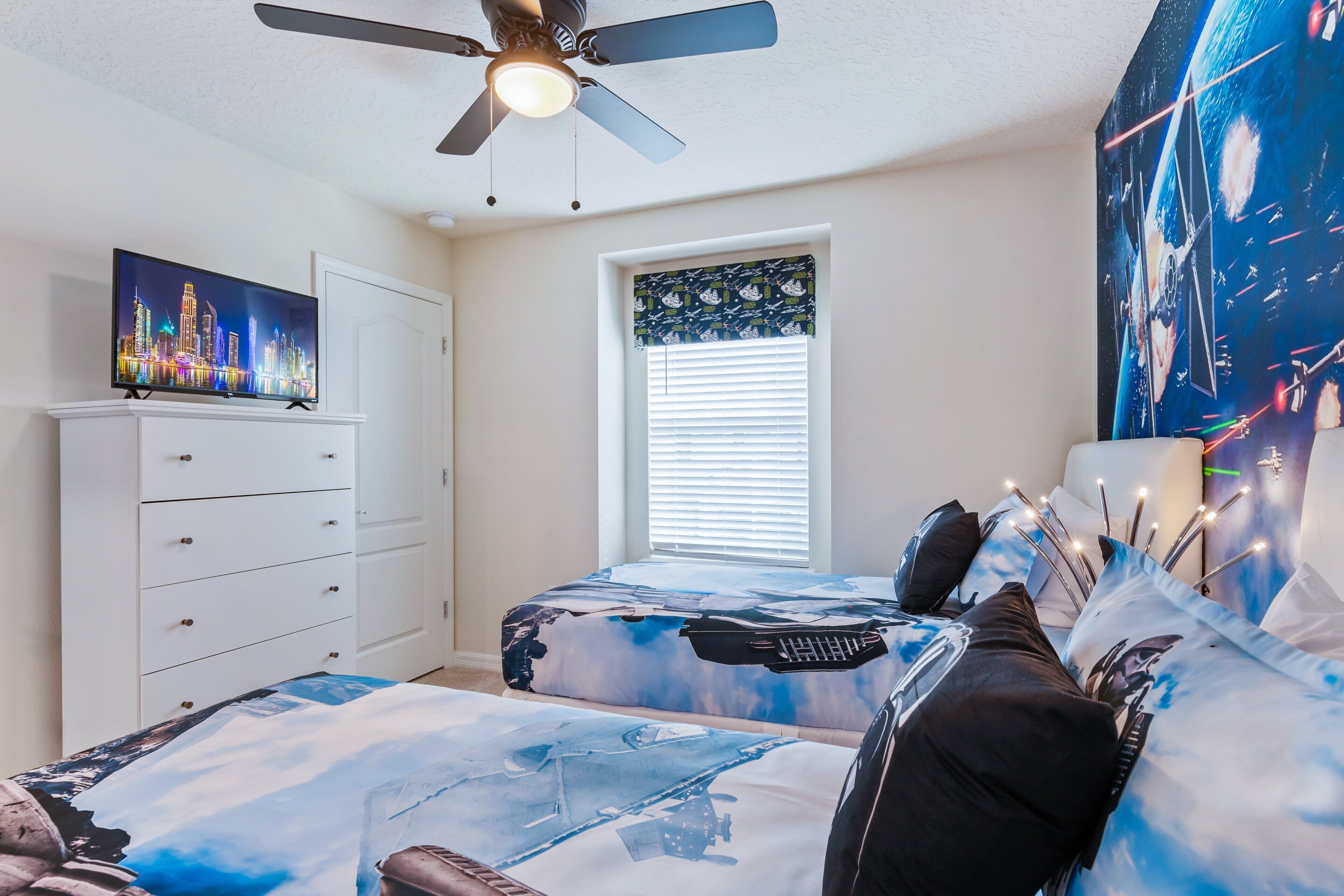 Une chambre avec deux lits, un ventilateur de plafond, un téléviseur posé sur une commode et une fenêtre avec des stores.