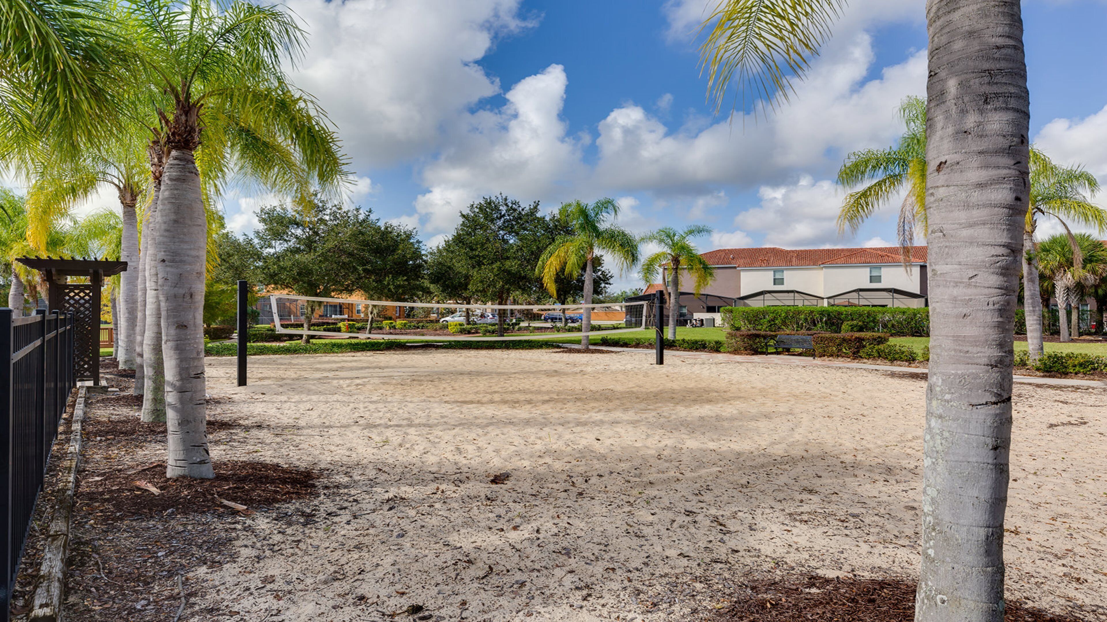 A sandy volleyball court surrounded by palm trees and a building with a red roof.