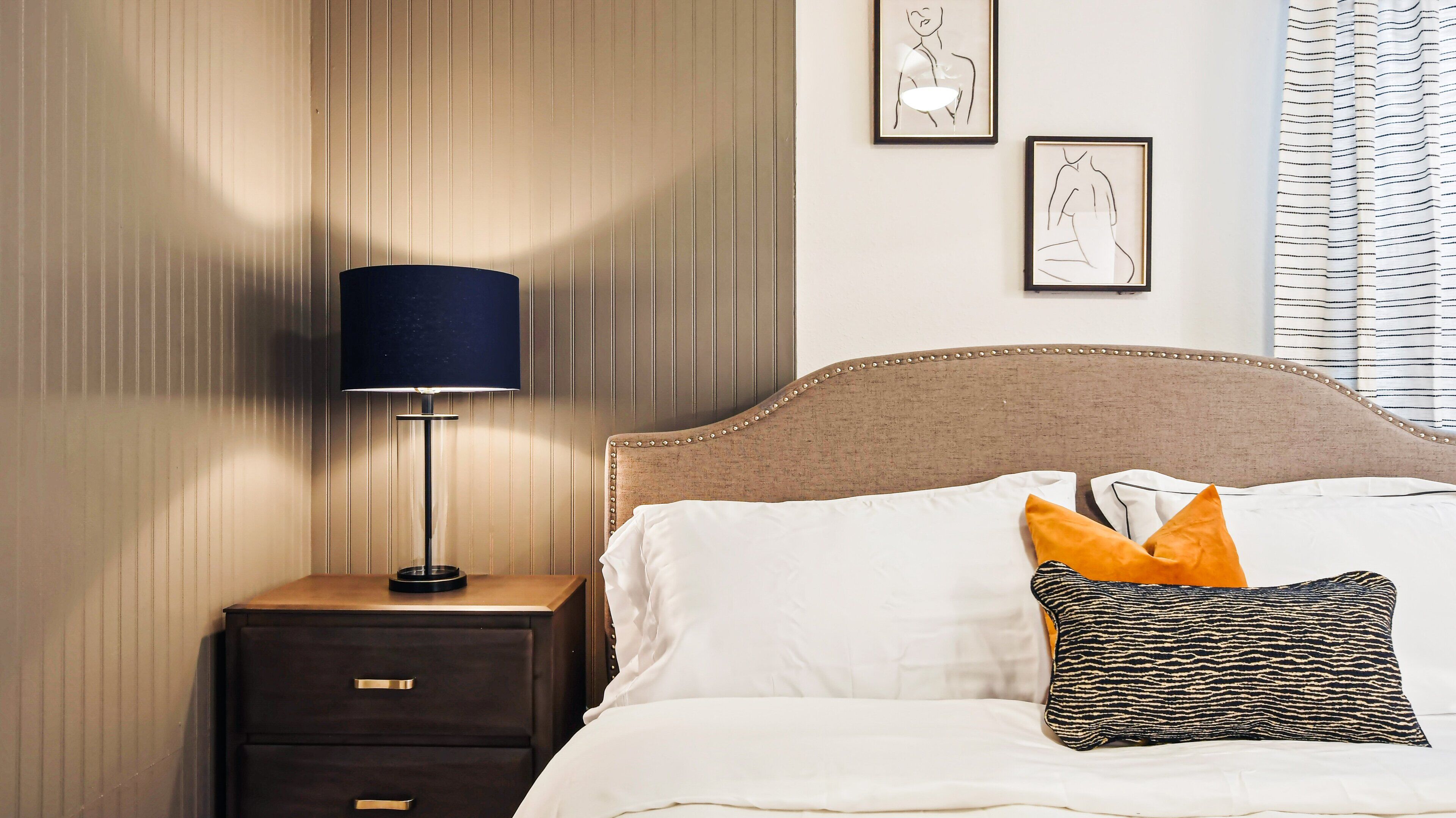 A neatly made bed with white and patterned pillows, a dark blue lamp on a wooden nightstand, and framed artwork on the wall.