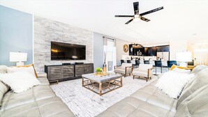 Un salon moderne doté d’un grand téléviseur à écran plat, d’un ventilateur de plafond et d’une cuisine en arrière-plan.