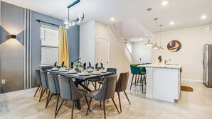 A modern kitchen with a central island, a dining area with a table set for four, and a staircase leading to an upper level. - Spacious Home Near Disney With Resort Amenities (Davenport)